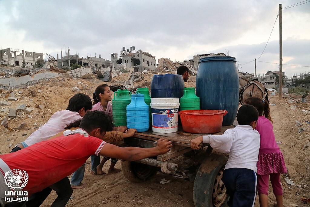 Palestinians in Gaza transporting water containers amid destroyed buildings in April 2025